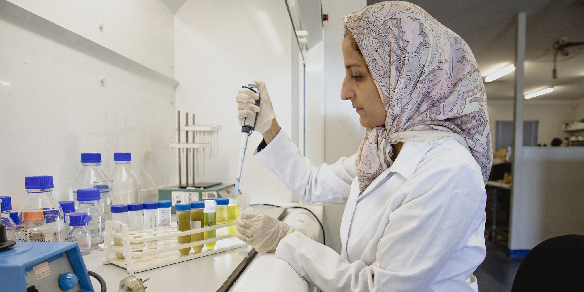 A worker in a lab coat and headscarf uses a pipette with test tubes, surrounded by bottles. The setting is focused and professional.