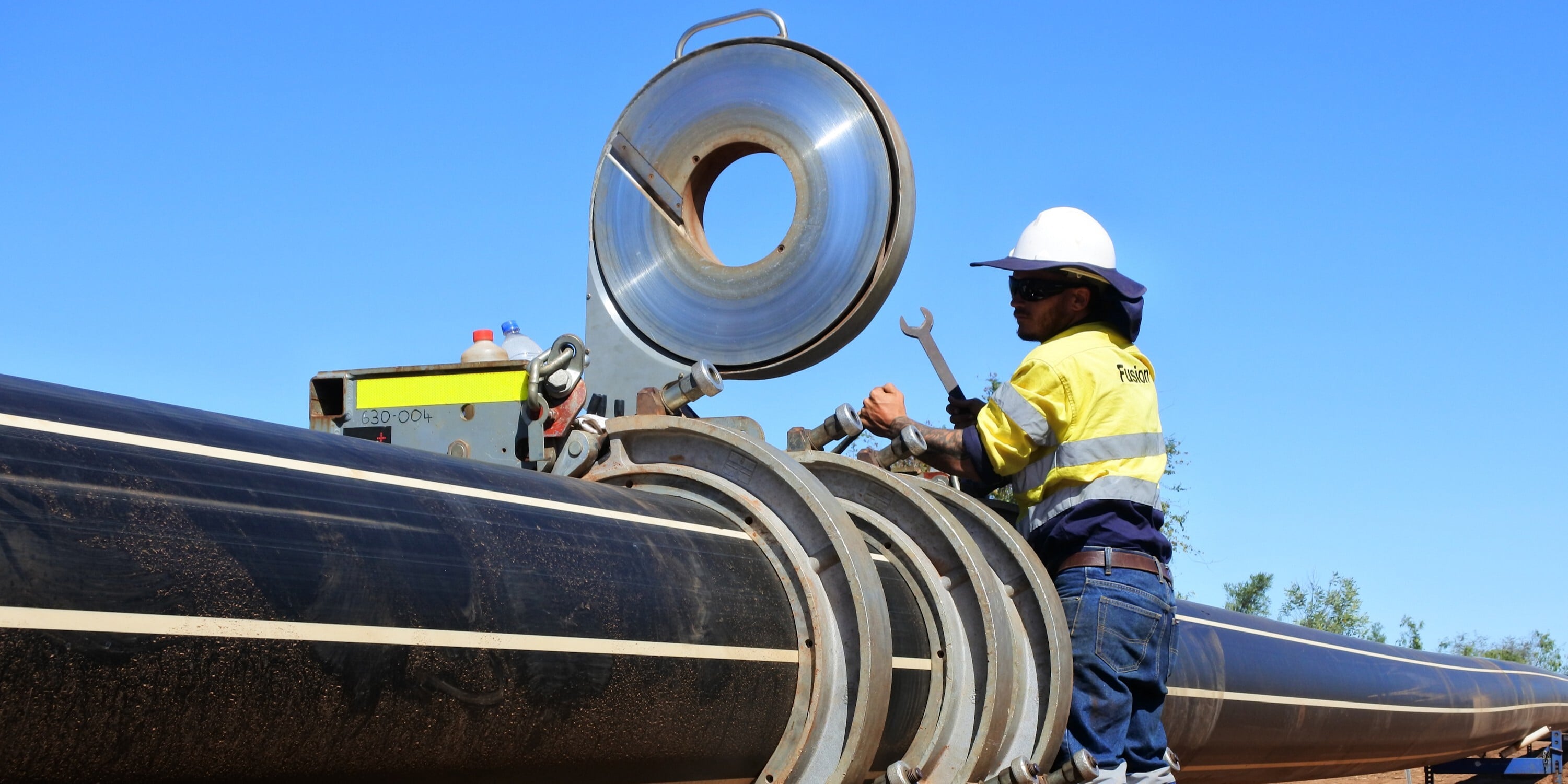 A man in safety gear, including a hard hat and glasses, is focused on repairing a pipe in an industrial setting.
