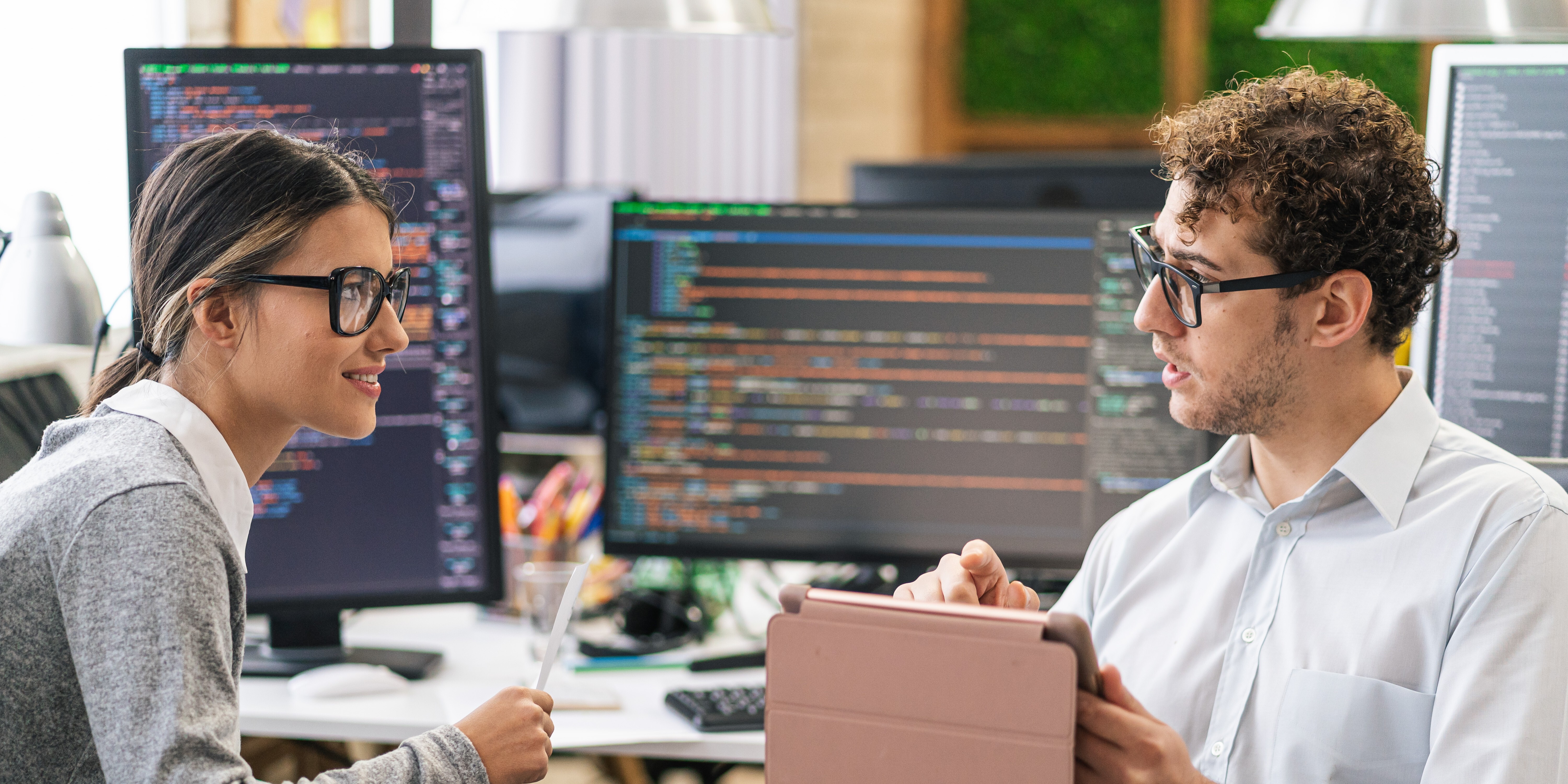 Two people collaborating on a tablet while seated in an office space, computers displaying code are in the background..