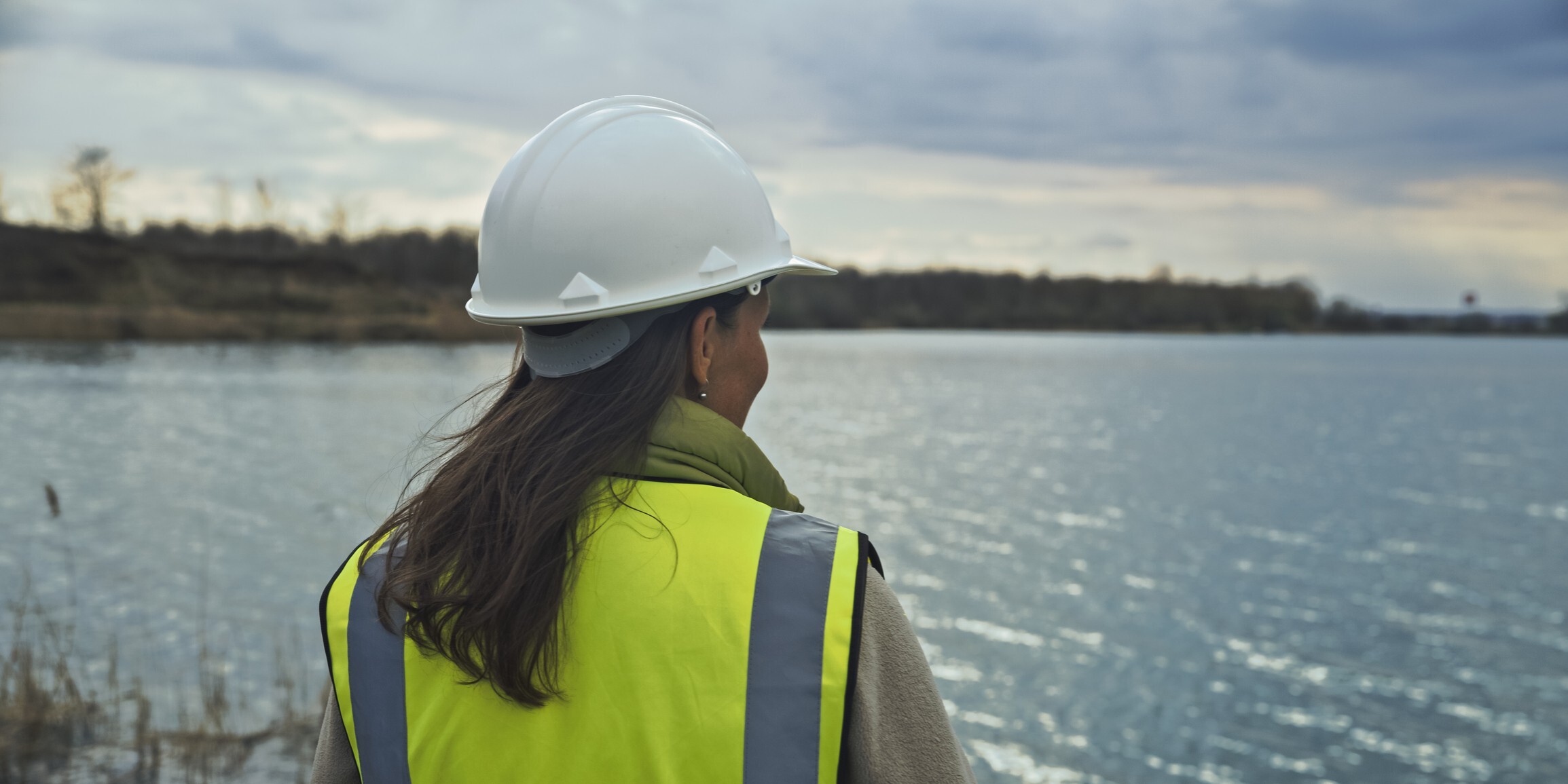 A woman wearing a hard hat stands by the water, observing the scene with a focused expression.
