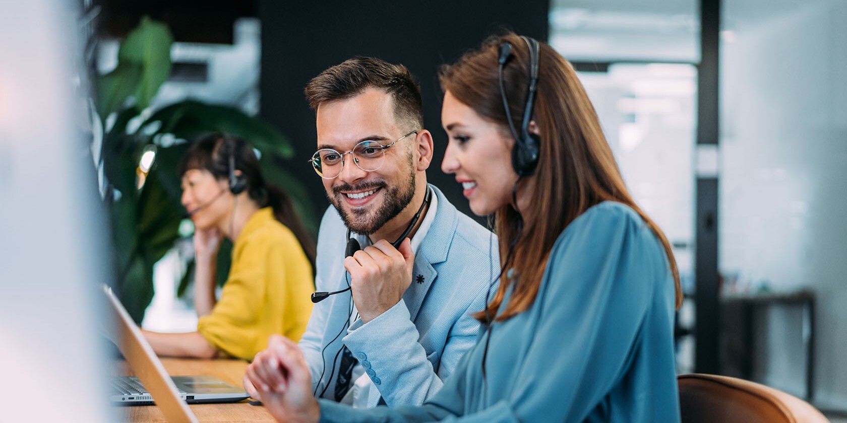 A team of three customer service agents wearing headsets works at desks with laptops. Two in the foreground are smiling and engaged in conversation.