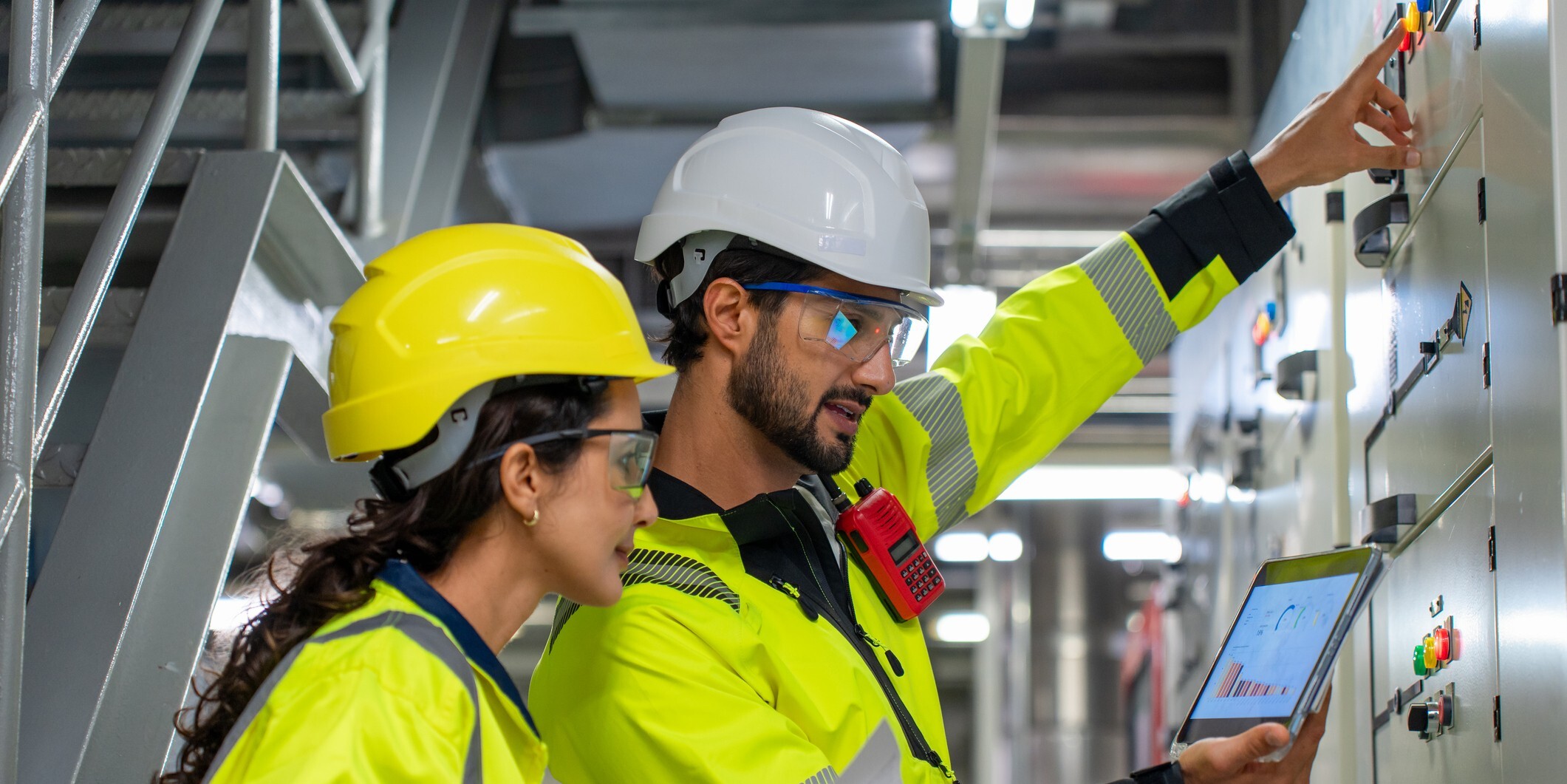 A male and female worker, both wearing safety gear, engage with an electrical control panel in an industrial setting.