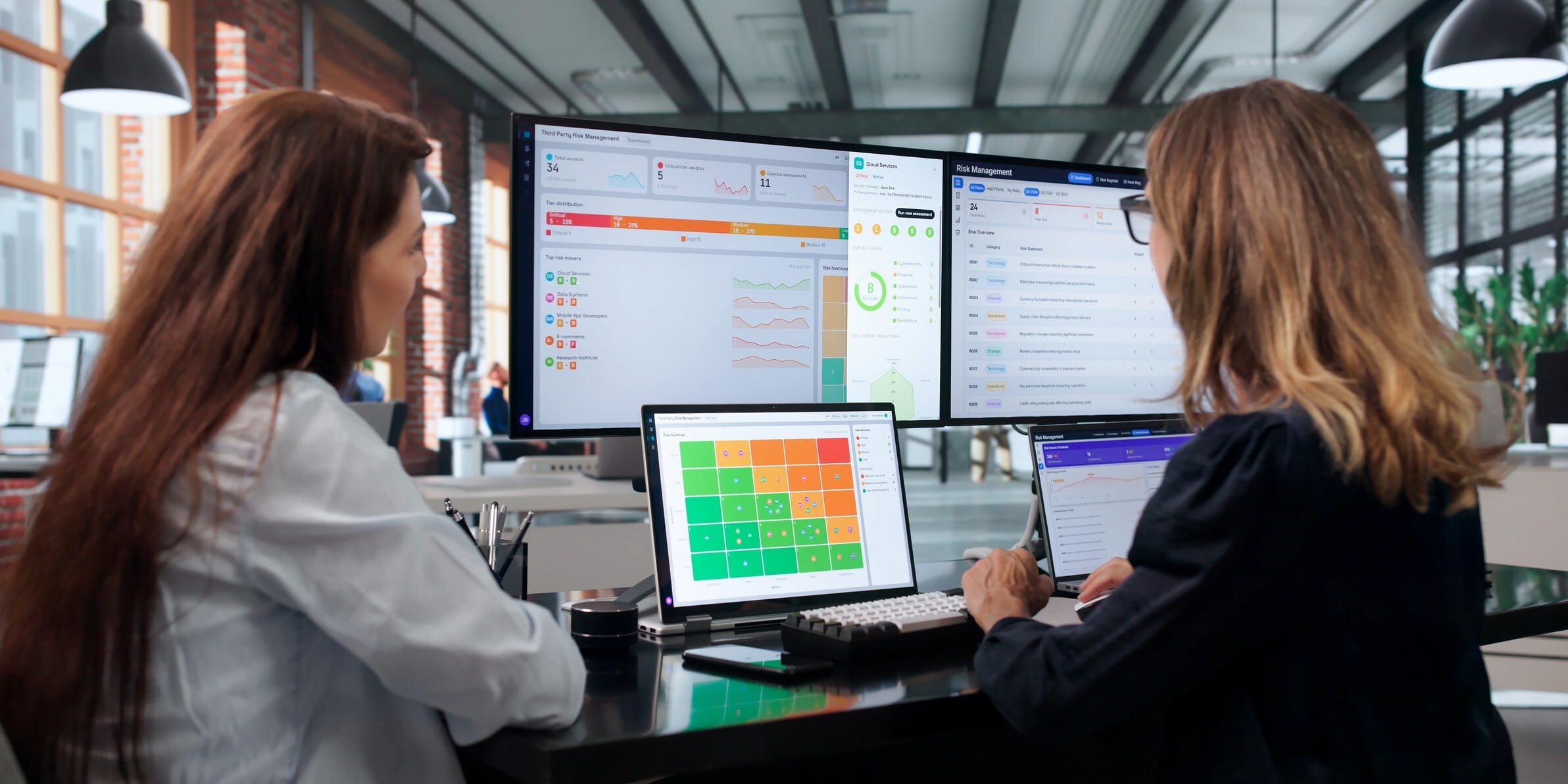 Two women seated at a desk, engaged in discussion while looking at a computer screen showing a red to green risk scale..