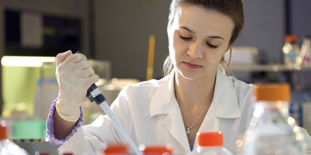 A scientist in a lab coat uses a pipette, focused on her work. Laboratory bottles with orange caps are in the foreground, creating an atmosphere of precision.