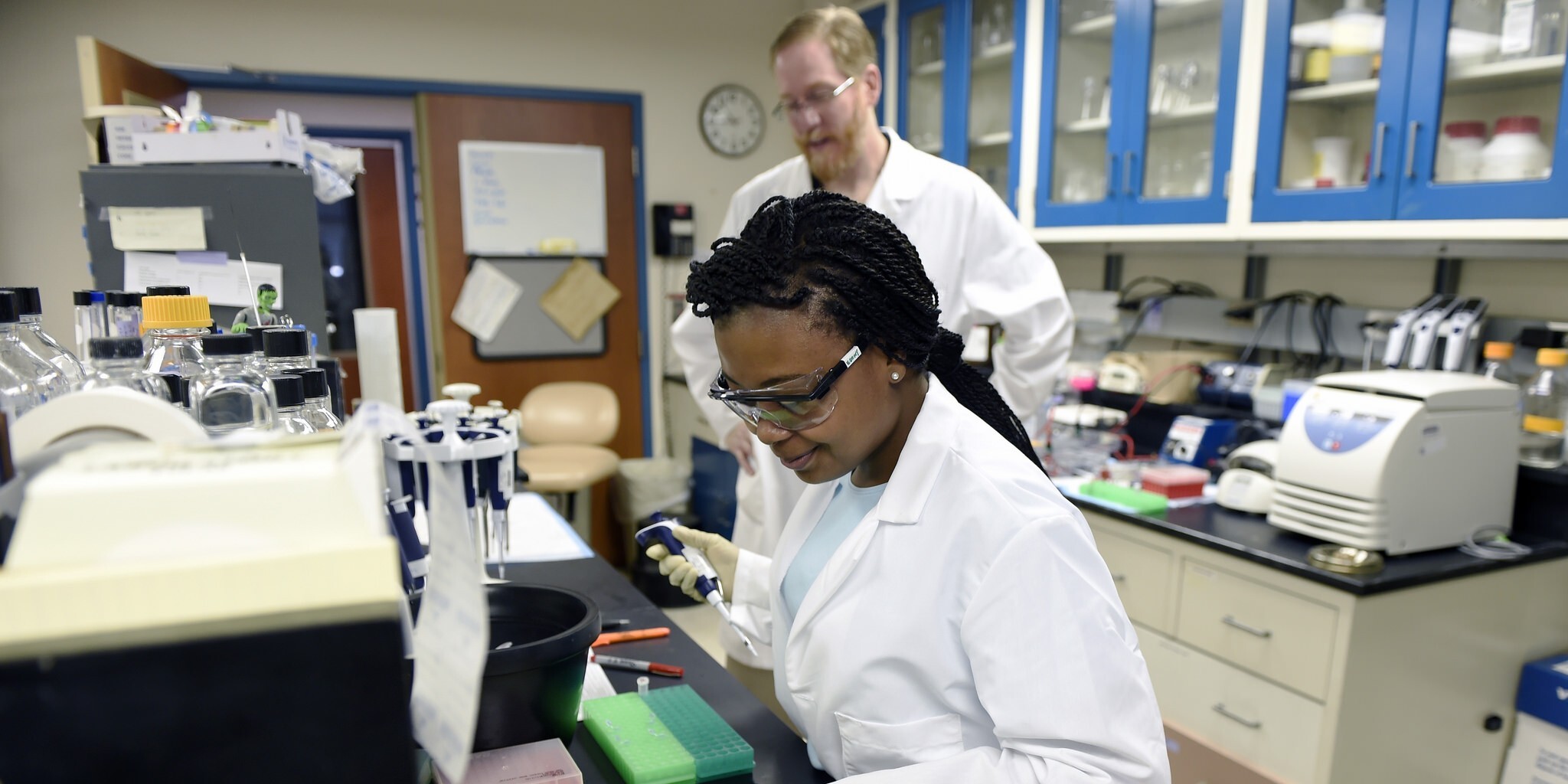 A scientist in a lab coat and safety goggles uses a pipette at a lab bench. Another person in a lab coat observes. The lab is equipped with scientific tools.