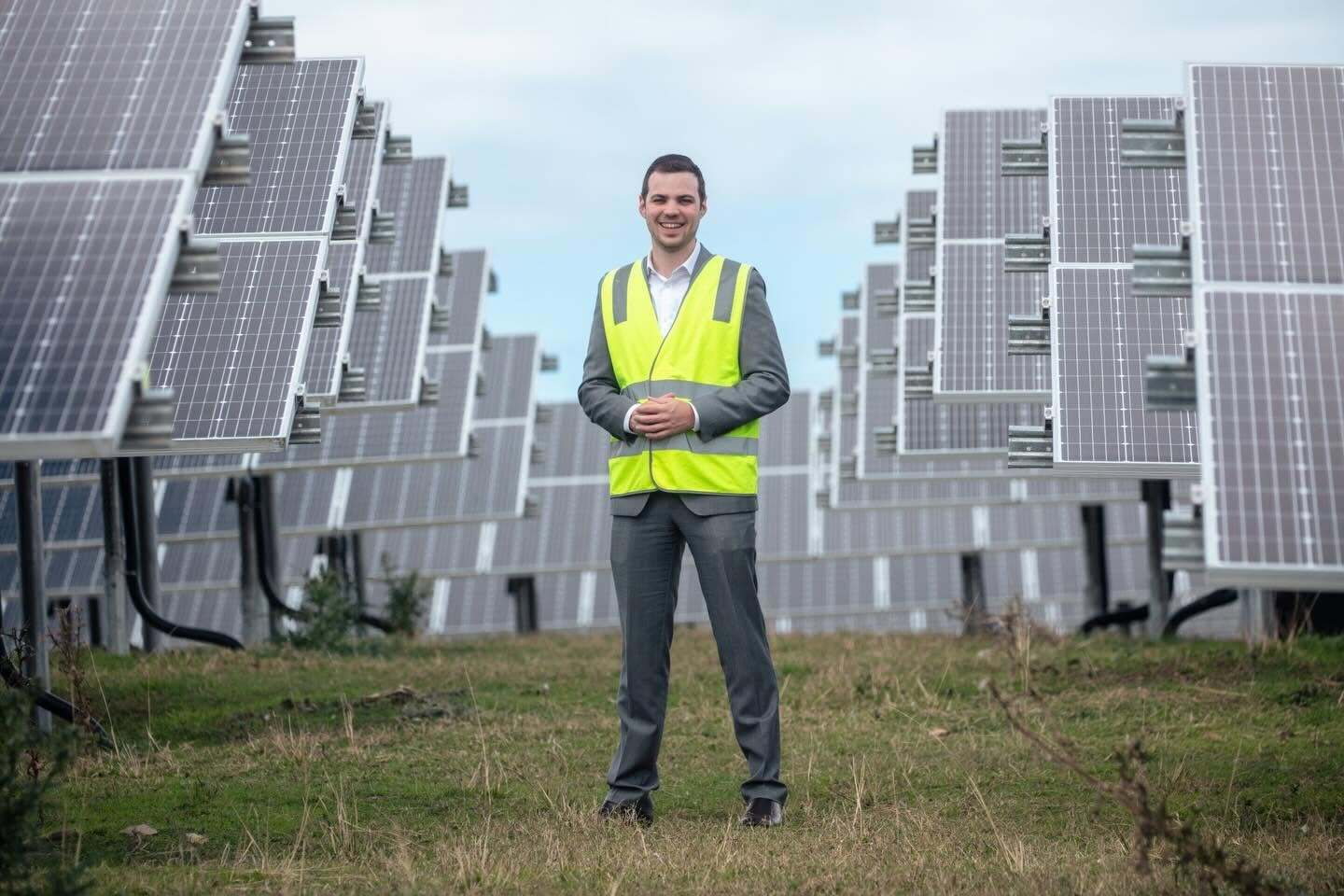 A person in a yellow high-vis vest stands on the grass in front of a solar farm.
