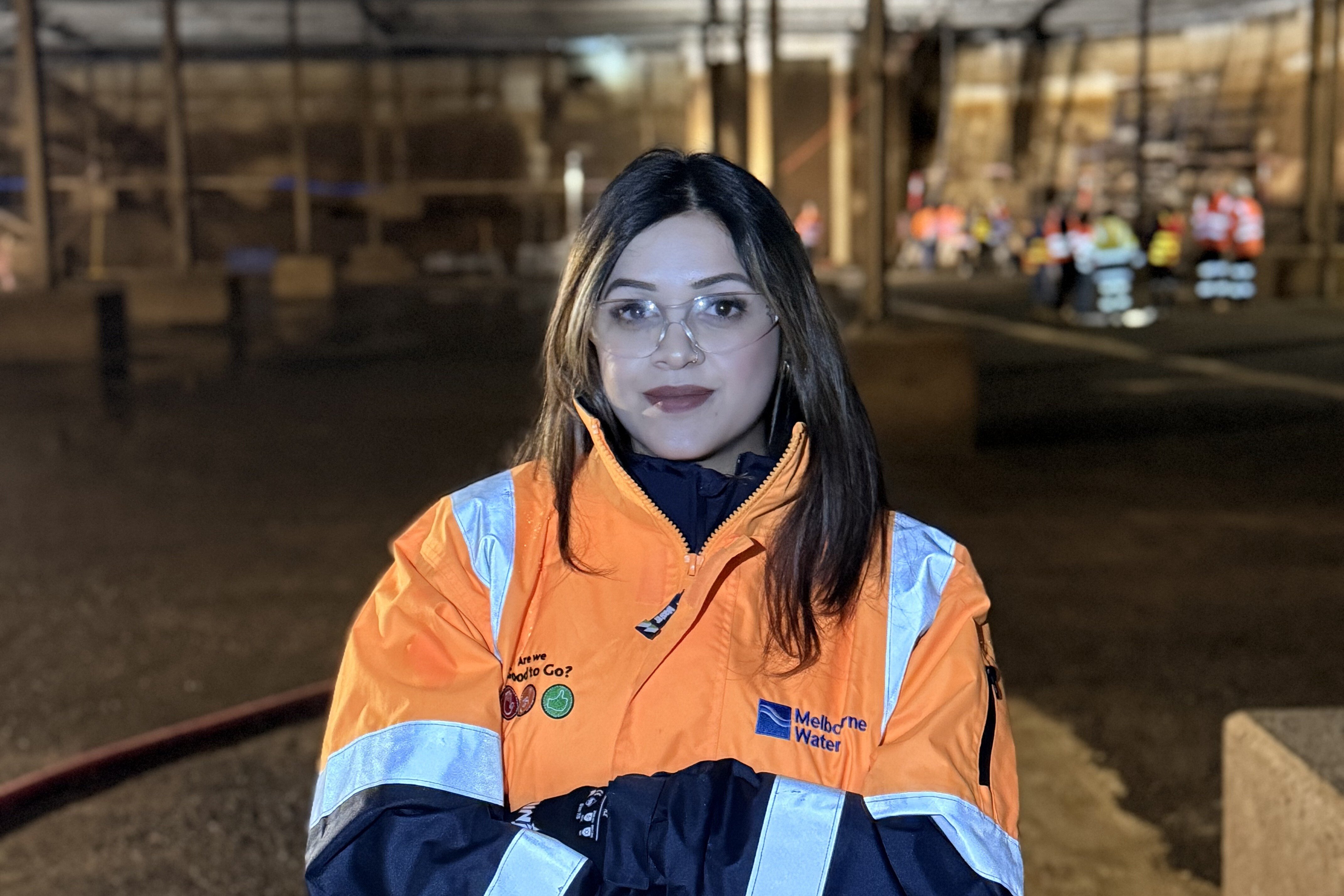 A person in an orange high-visibility jacket stands in a dimly lit industrial setting, with a serious expression, surrounded by distant workers.