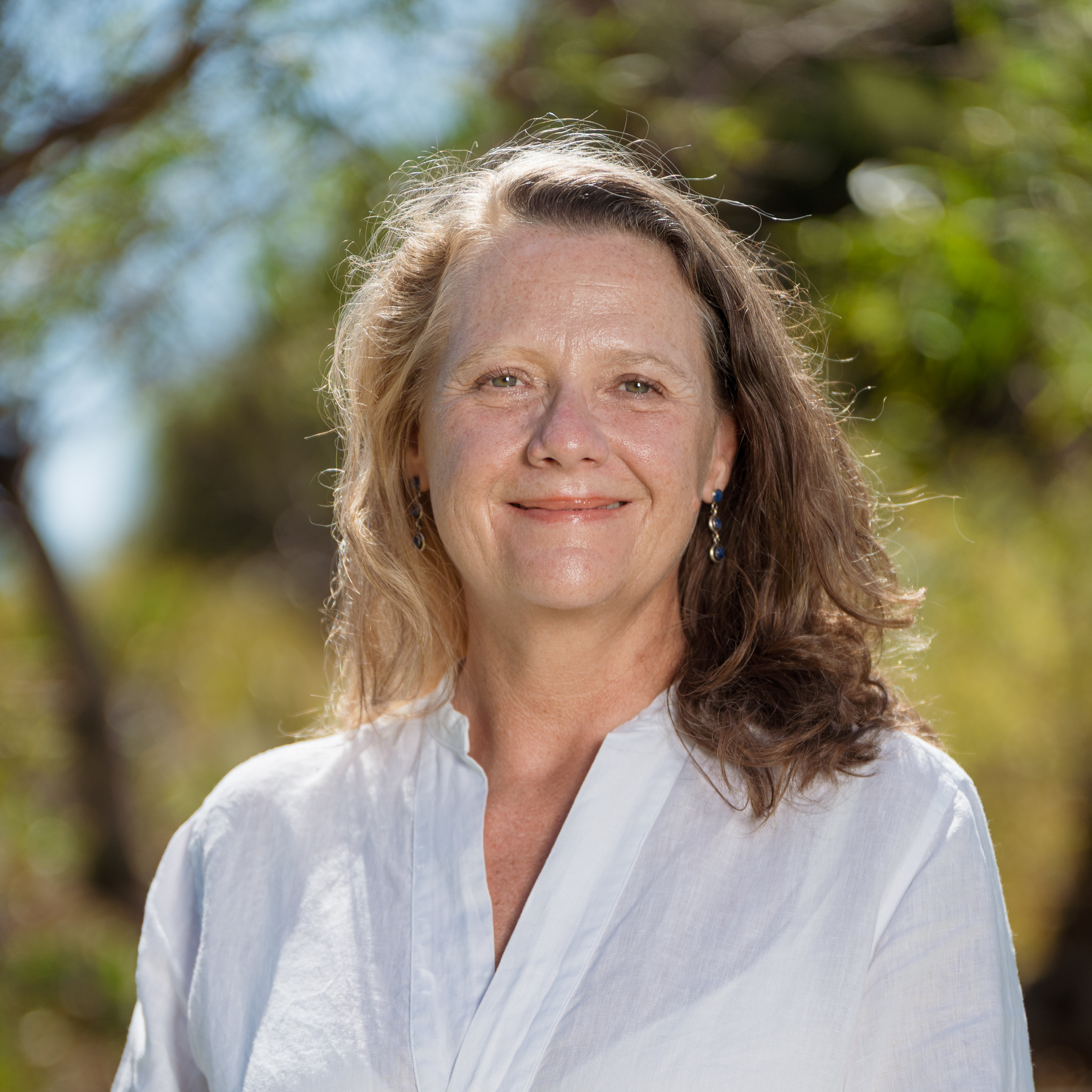 Smiling woman with long hair wearing a white blouse stands outdoors. Background features blurred greenery and sunlight, conveying a warm, cheerful tone.