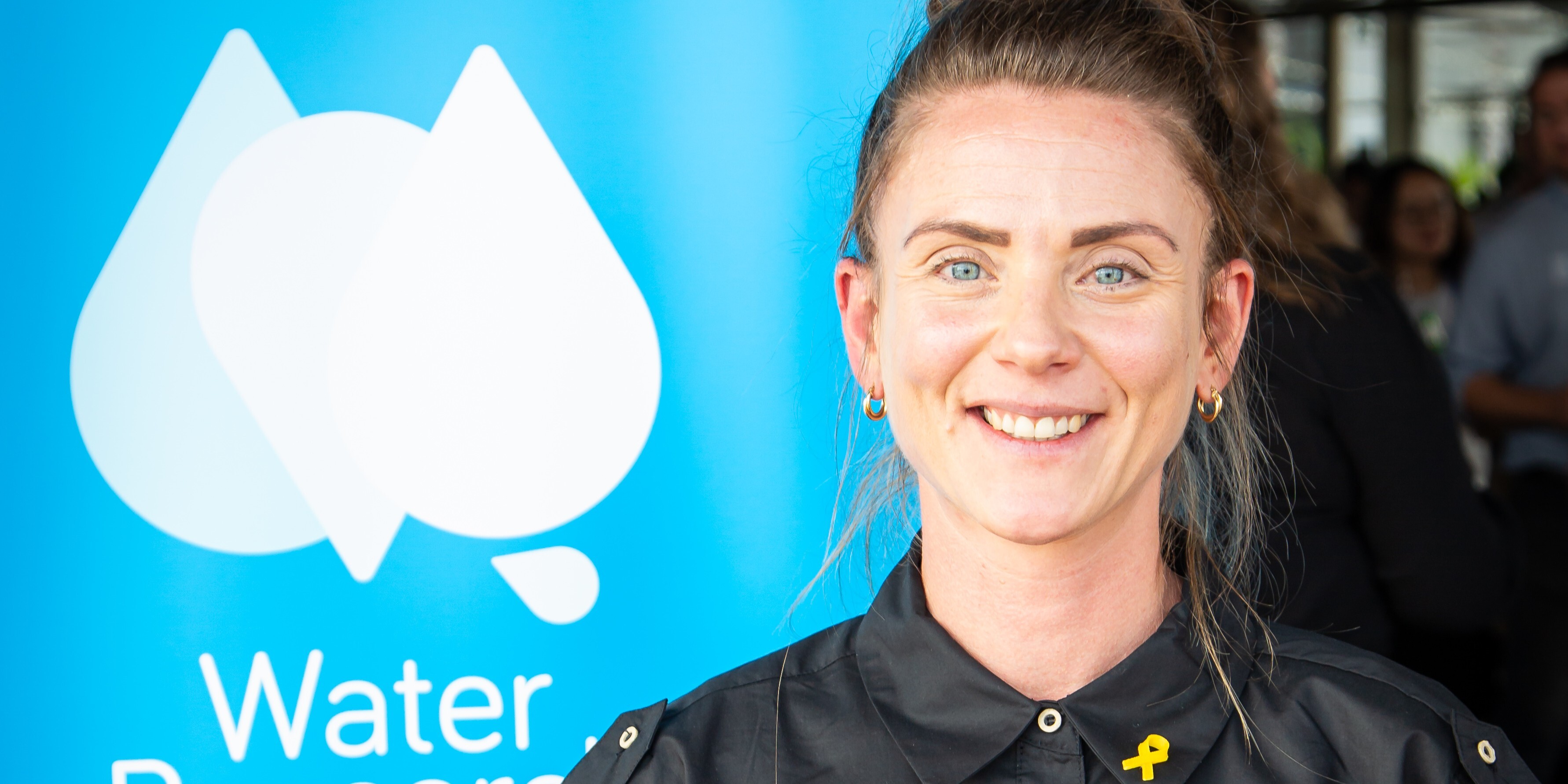 Ortal with a yellow ribbon pin on her shirt stands in front of a blue 