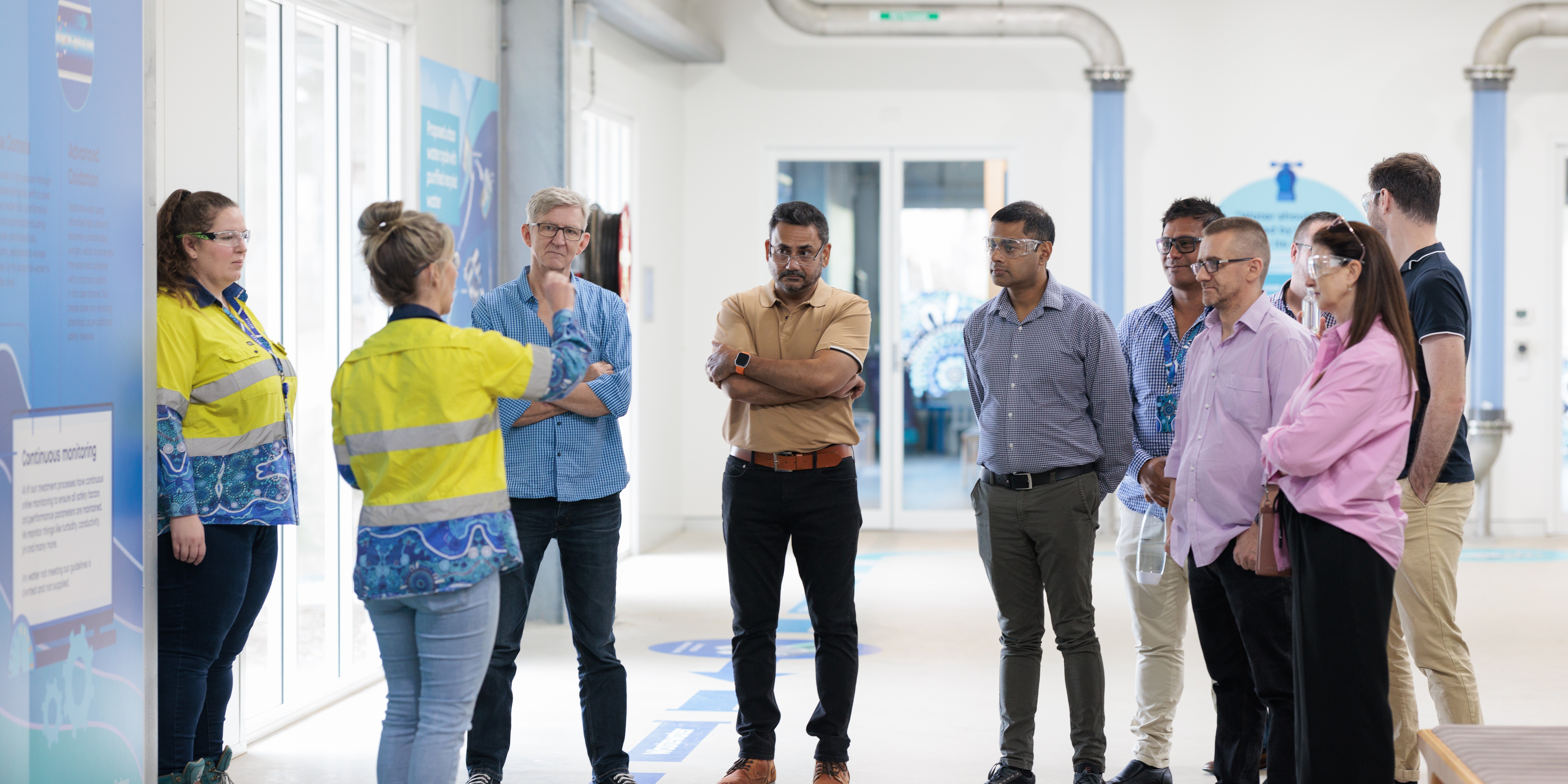 A group of people stand in a well-lit industrial space, attentively listening to a person wearing a yellow safety vest pointing at a display. Photo courtesy of Sydney Water.