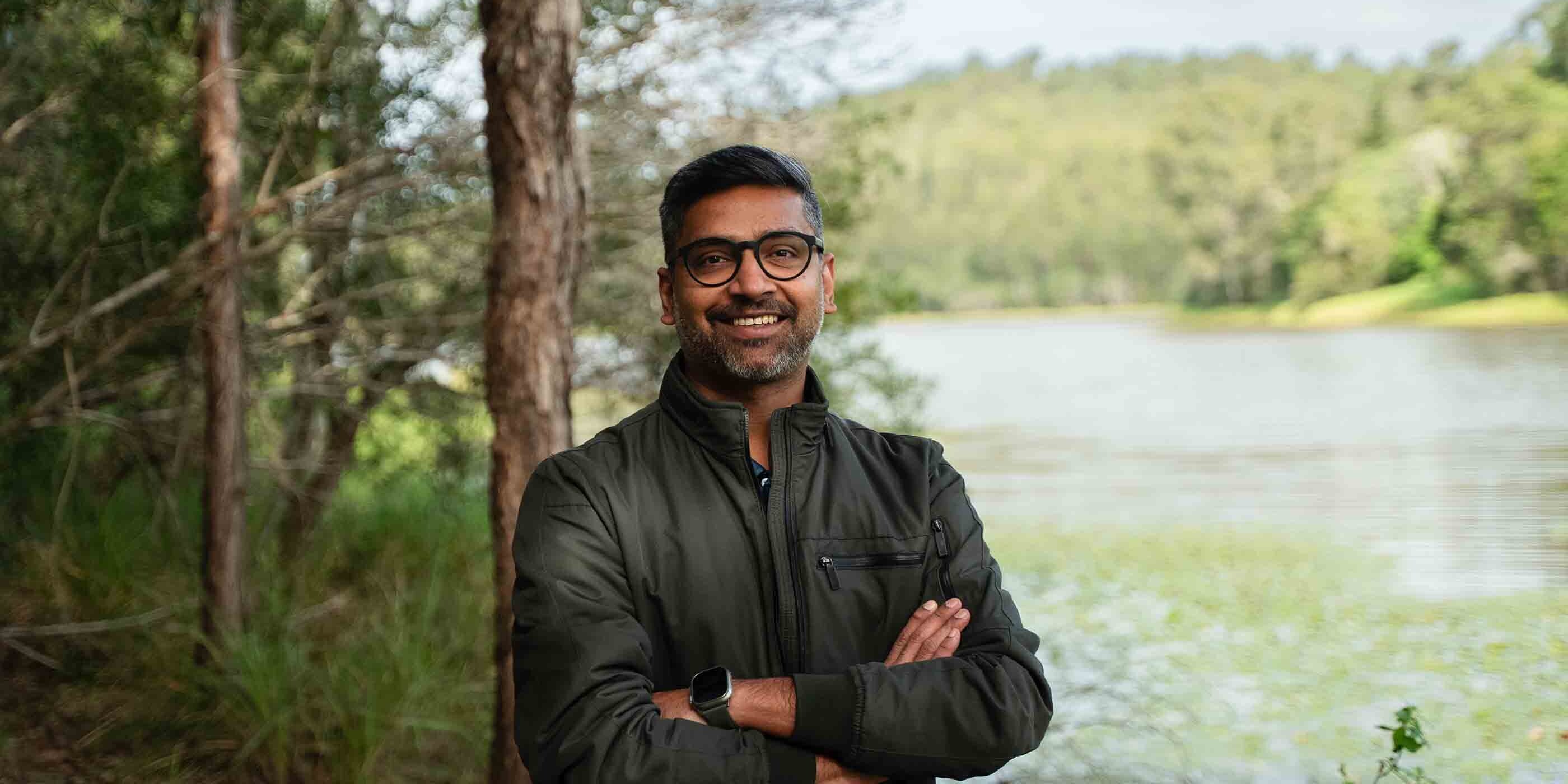 Smiling man with glasses and a dark jacket stands confidently with arms crossed in front of a lake, surrounded by lush green trees.
